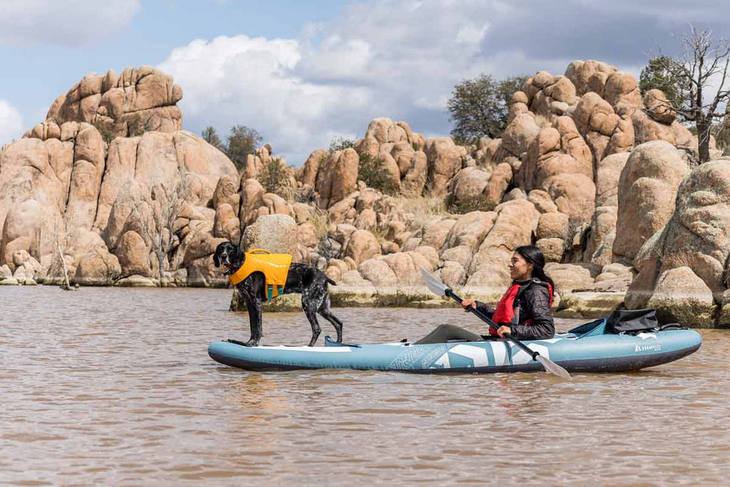 Woman kayaks in an inflatable kayak with a black lab dog standing on the bow of the boat.