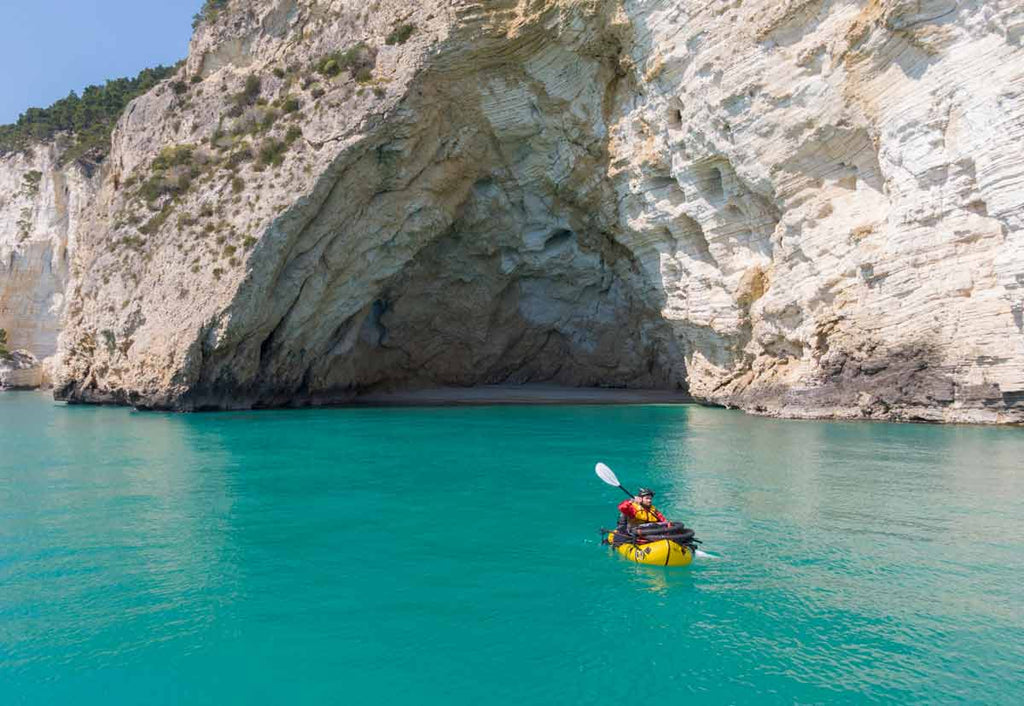 Man packrafts across turquoise water near a natural rock formation on a clear and sunny day